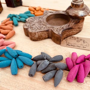 Op de afbeelding: Een houten wierookbrander met een handvormig ontwerp en een kleine kom om wierookkegels te verbranden. De brander is omringd door een verscheidenheid aan kleurrijke wierookkegels, waaronder roze, blauw, grijs en oranje.