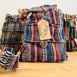 May include: Three woven fabric backpacks featuring vertical stripes in red, blue, black, and white. Each bag has a front pocket with a wooden button closure and a brown paper tag attached with string that reads The Happy Hippie Bus.