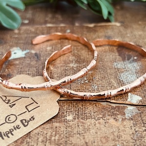 Op de afbeelding: Twee koperen armbanden met een gestructureerd bamboe-ontwerp. De armbanden zijn open en hebben een gepolijste, metalen afwerking. Een beige papieren label met de gedrukte tekst Happy Hippie Bus is onder de armbanden zichtbaar.