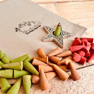 Pode incluir: Uma variedade de cones de incenso em tons de verde, laranja e vermelho, juntamente com um porta-incenso em forma de estrela. Um envelope de papel castanho com um logótipo está no fundo. Os artigos estão dispostos numa superfície de madeira clara.