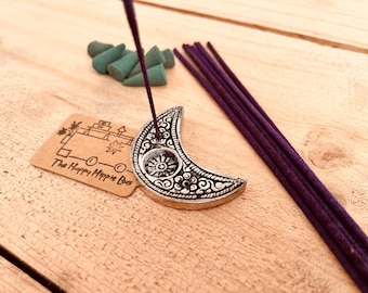 Könnte beinhalten: Ein silberner Halbmond-Räucherstäbchenhalter mit filigranen Verzierungen. Der Halter steht auf einer Holzoberfläche mit einem violetten Räucherstäbchen und einem kleinen Stapel grüner Räucherkegel.