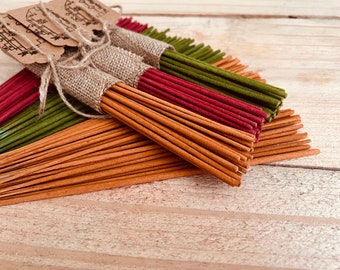 May include: Bundles of incense sticks in shades of brown, green, and red are displayed on a light wooden surface. Some bundles are wrapped in burlap and tied with twine, with small tags attached. The incense sticks are arranged in a fan-like pattern.