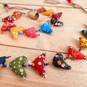 May include: A colourful necklace made of fabric hearts with different patterns and beads. The necklace is strung on a brown cord and has a bell charm.