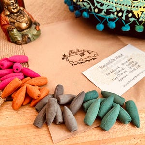 Puede incluir: Una variedad de conos de incienso en rosa, naranja, gris y verde, dispuestos sobre una superficie de madera. Una tarjeta indica "Tranquility Blend" con descripciones de aromas. Una pequeña estatua de Buda y una tela estampada están en el fondo.