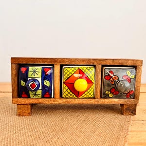 May include: A wooden spice rack with three ceramic drawers. Each drawer is square and features a unique hand-painted design with colourful patterns and knobs. The rack is made of light brown wood and has small feet.