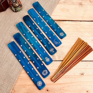 Könnte beinhalten: Eine Sammlung blauer Räucherstäbchenhalter aus Holz mit weißen Sternmustern und verschiedenen Himmelsymbolen. Ein Bündel Räucherstäbchen ist ebenfalls vorhanden. Die Halter sind auf einer Holzoberfläche angeordnet.