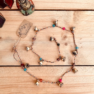 Puede incluir: Un collar decorativo con pequeñas campanas doradas y cuentas de colores. El collar está hecho de hilos trenzados marrones, rojos y negros. El collar está sobre una superficie de madera.