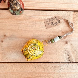May include: A yellow bell-shaped ornament with white and green floral designs. It has a gold ring, a white and green bead, and a natural fibre string. A small tag with text is attached.