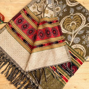 Op de afbeelding: Een gevouwen textiel met een beige, rode en olijfgroene kleurstelling. Het textiel heeft ingewikkelde patronen, waaronder bloemen en geometrische ontwerpen. De randen zijn afgewerkt met franjes.