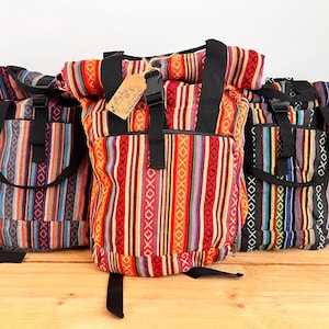 May include: Three roll-top backpacks featuring vibrant, multi-coloured woven fabric with vertical stripes and geometric patterns. Each bag includes black nylon straps, a plastic buckle closure, and a small rectangular paper tag attached by twine.