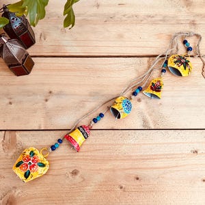 May include: A decorative garland of yellow metal bells and a heart-shaped ornament, hand-painted with floral designs in red, orange, and blue. The bells are strung together with a natural twine and blue beads. The background is a light brown wooden surface.