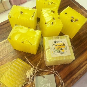 May include: Yellow, square-shaped soaps with dried flower accents are arranged on a wooden cutting board. One soap is wrapped with a label that reads "Venice Chamomile Tea." A tag with the text "Twenty Twenty by Vesta" is also present.