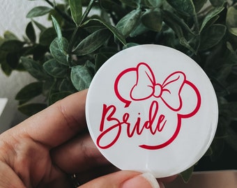 May include: A white round button with a red Minnie Mouse bow and the word "Bride" in red script. The button is held in front of a background of green leaves.