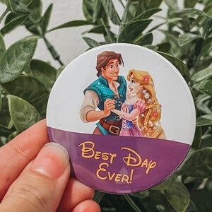 Op de afbeelding: Een ronde button met een cartoon illustratie van een stel en de tekst "Best Day Ever!" in gouden letters op een paarse achtergrond. De bovenste helft van de button toont een man en een vrouw in een romantische pose.