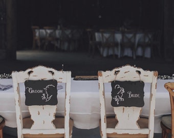 Puede incluir: Dos sillas blancas envejecidas con asientos de arpillera y letreros de pizarra negros que dicen "Groom" y "Bride". Los letreros presentan ilustraciones de siluetas. Las sillas están colocadas en una mesa con un mantel blanco, en un entorno rústico.