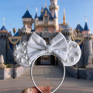 May include: A silver headband with grey mouse ears and a matching bow. The ears are decorated with white Mickey Mouse head shapes. The background features a castle under a clear blue sky.