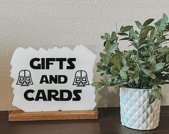 Könnte beinhalten: Ein klarer Acrylschild mit den Worten "GIFTS AND CARDS" in fetter schwarzer Schrift, zusammen mit zwei Darth-Vader-Helm-Grafiken. Das Schild ist auf einem Holzsockel montiert. Rechts steht eine Topfpflanze mit grünen Blättern.