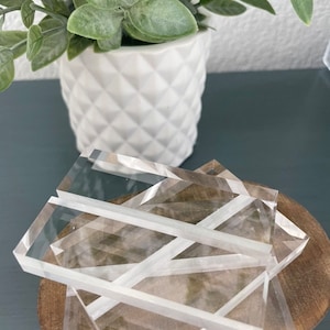 May include: Clear acrylic coasters on a wooden base. The rectangular, transparent coasters have a frosted look. A white, diamond-patterned pot with green foliage is in the background. The coasters are ideal for protecting surfaces.