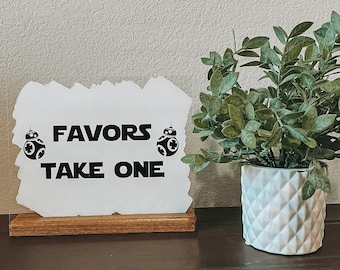 Könnte beinhalten: Ein weißes Schild mit dem Text "FAVORS TAKE ONE" in Schwarz, mit zwei BB-8-Droiden-Grafiken. Das Schild ist auf einem Holzsockel montiert. Rechts befindet sich ein weißer geometrischer Topf mit einer grünen Pflanze.