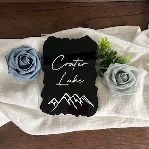 Op de afbeelding: Een zwart acrylbord met witte tekst "Crater Lake" en een bergafbeelding. Het bord wordt geflankeerd door blauwe en grijze stoffen rozen en groen op een witte doek.