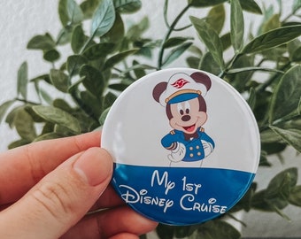 May include: A round button with a white top half featuring Mickey Mouse in a captain's uniform. The bottom half is blue with the text "My 1st Disney Cruise" in white script. The button is held in front of a green leafy plant.