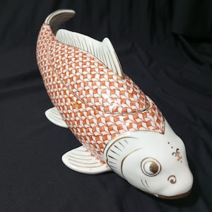 Puede incluir: Una figura de pez koi de cerámica con tapa. El pez es blanco con patrones de escamas naranjas y blancas. Las aletas y la cola son blancas con detalles dorados. El pez tiene una cara detallada con detalles dorados y una boca curva.