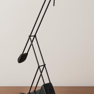 Può includere: Una lampada da scrivania nera dal design geometrico. La lampada presenta una base rettangolare, supporti angolati e una testa regolabile. La testa della lampada è di forma ovale e piatta. La lampada è in metallo con finitura opaca.