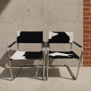 May include: Two modern chairs with black and white cowhide seats and backs. The chairs have a chrome frame and black armrests. The chairs are positioned in front of a concrete wall and a brick wall.