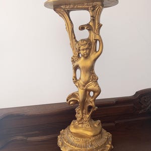 May include: A gold-colored decorative stand with a cherub figure. The stand has a round top and a detailed base with a floral design. The cherub is holding a floral element. The stand is made of a metallic material.