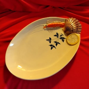 Puede incluir: Plato ovalado para servir con superficie color crema. Presenta una langosta naranja en relieve, una concha marrón, una rodaja de limón amarilla y detalles de hojas negras. El plato está sobre un fondo rojo.