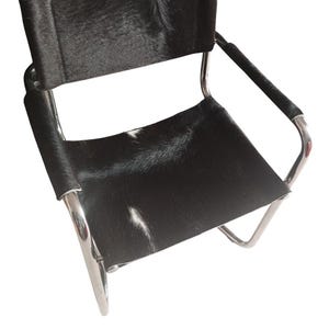Könnte beinhalten: Moderner Stuhl mit Sitzfläche und Rückenlehne aus schwarz-weißem Kuhfell. Der Stuhl hat ein verchromtes Metallgestell und Armlehnen und bietet ein schlankes, minimalistisches Design. Geeignet für Wohn- oder Büroräume.