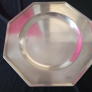 May include: A silver-colored, octagonal plate with a raised rim and a circular indentation in the center. The plate has a polished finish, reflecting the surrounding environment. The plate is likely made of metal and is designed for serving food.
