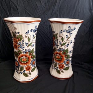Op de afbeelding: Twee witte keramische vazen met een blauw en oranje bloemenmotief. De vazen hebben een geribbeld ontwerp en een zeshoekige basis.