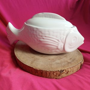 以下が含まれることがあります： 蓋付きの白い陶器製の魚の形をしたサービングディッシュ。ディッシュは木製の台座の上に置かれています