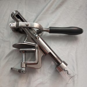 May include: A silver metal wine bottle opener with a black handle and a clear plastic corkscrew. The opener has a clamp to hold the bottle in place.
