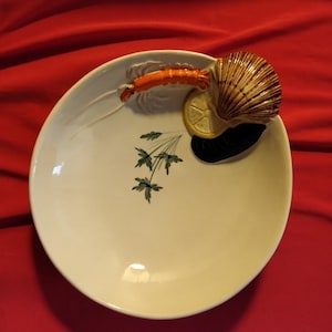 Pode incluir: Um prato de servir de cerâmica branca com um desenho em relevo de um camarão, uma fatia de limão e uma concha de vieira. O prato tem um desenho de salsa verde.