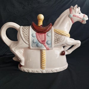 Könnte beinhalten: Eine weiße Keramik-Teekanne in Form eines Karussells. Das Pferd hat einen braunen Sattel, einen gelb-weiß gestreiften Pfosten und ein rosafarbenes und blaues Blumenmuster an der Seite.