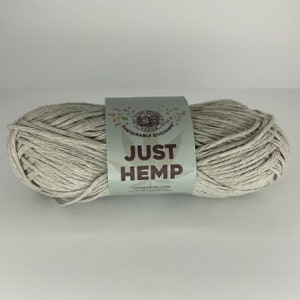 May include: A skein of Lion Brand Just Hemp yarn in a natural, light beige color. The label features the text "JUST HEMP" in a bold font, with the Lion Brand logo and the words "Sustainable Stitching".