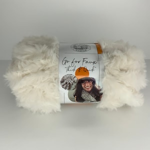 May include: A skein of white faux fur yarn with a label that reads "Go for Faux! Thick & Quick" and features a woman wearing a scarf made from the yarn.