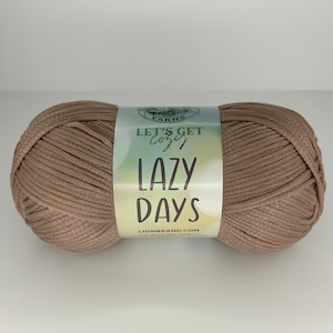 May include: A large ball of tan yarn with a label that reads "Let's Get Cozy Lazy Days." The yarn is tightly wound and appears soft. The label has a green and white watercolor design.