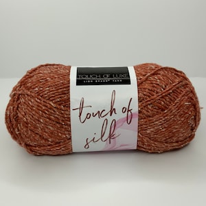 May include: A skein of Touch of Luxe yarn in the color "Terracotta" by Lion Brand Yarn. The yarn is a blend of acrylic and silk and has a speckled texture.