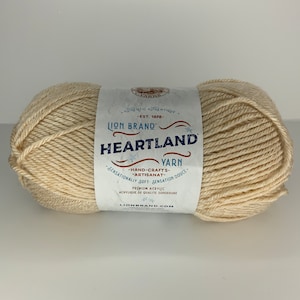 May include: A skein of Lion Brand Heartland yarn in a cream color. The label reads "Lion Brand Heartland Yarn - Hand-Crafts - Artisanat - Sensationally Soft Sensation Douce - Premium Acrylic - Acrylique de Qualite Superieure - LionBrand.com".