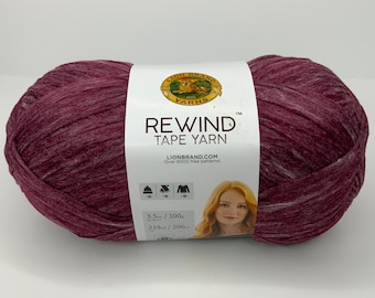 Può includere: Un gomitolo di filato Lion Brand Rewind Tape Yarn di colore bordeaux con un'etichetta bianca che dice "Lion Brand Yarns Rewind Tape Yarn" e "Lionbrand.com Oltre 8000 modelli gratuiti!" L'etichetta include anche il peso del filato, 100g / 3.5oz, e il metraggio, 200m / 219yd.