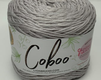 May include: A skein of light gray Coboo yarn. The label features the brand name and the text "Over 8000 free patterns!" and "LIONBRAND.COM". The yarn is tightly wound, showing the texture of the fibers. The label also has a pink ribbon.