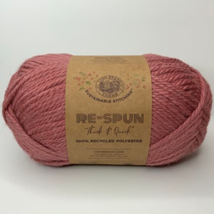 Op de afbeelding: Een bol roze garen met het label "RE-SPUN Thick & Quick 100% RECYCLED POLYESTER", het Lion Brand Yarns logo en de tekst "Sustainable Stitching".