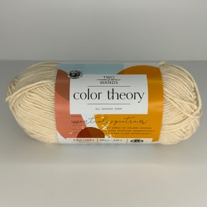 Puede incluir: Una madeja de hilo de color crema etiquetada como "Two of Wands Color Theory Essential Spectrum All Season Yarn". La etiqueta presenta un diseño abstracto colorido y el texto "Una gama de colores elegidos para trabajar juntos armoniosamente o para destacar por sí solos sin esfuerzo". El hilo es de 3,5 oz/100 g y 246 yds/225 m.