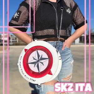 May include: A white, round handbag with a compass design in the center, featuring red and gray accents. The bag has a silver chain strap and the text "YOU MAKE STRAY KIDS STAY" around the edge. The bag is worn with a black mesh top and ripped denim jeans.