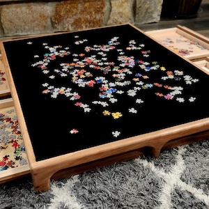 May include: A wooden puzzle table with a black surface, surrounded by four pull-out trays. The table is covered with colorful puzzle pieces. The trays contain sorted puzzle pieces. The table is on a gray rug.