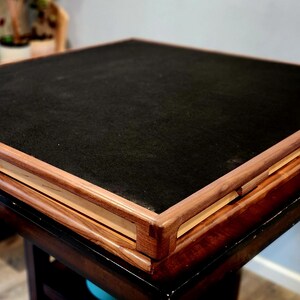 May include: A square wooden table with a dark, felt-like surface. The table has a dark brown base and a lighter wood frame around the top. The table is approximately 76 cm square.
