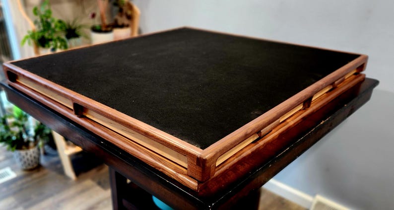 May include: A square wooden table with a dark, felt-like surface. The table has a dark brown base and a lighter wood frame around the top. The table is approximately 76 cm square.